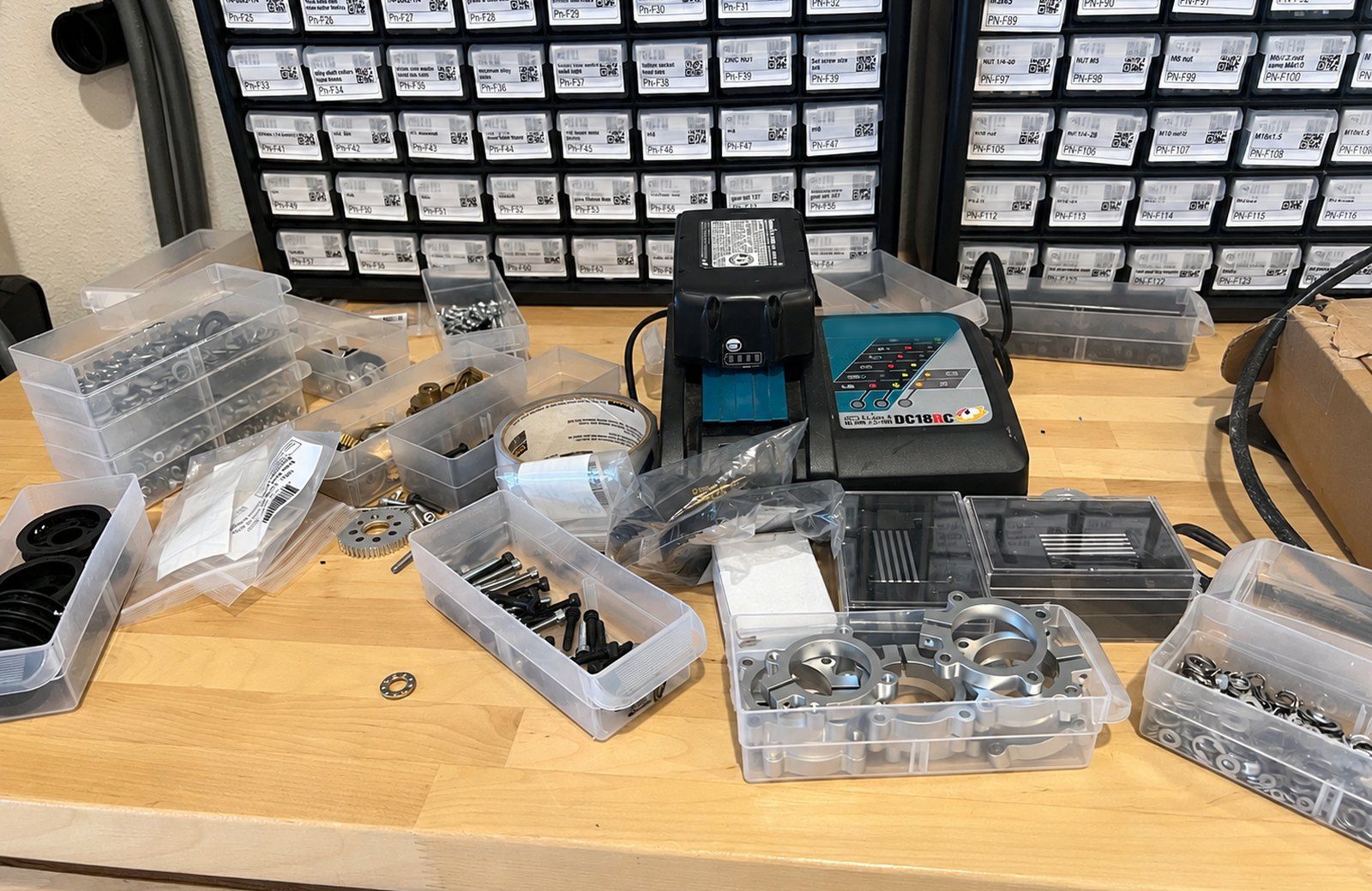 A real workbench: a labelled parts cabinet, a battery charger, tape, screws, gears and aluminum housings